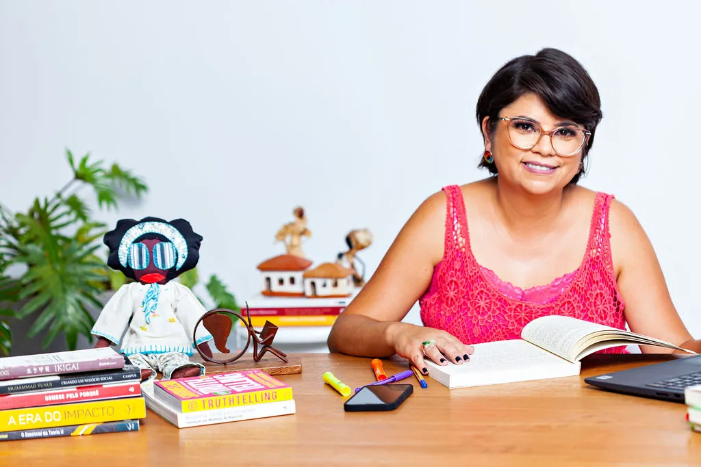 mulher sorrindo em mesa com livros e uma boneca de pano, é a Andreia Duarte fundadora da Ideia. Ela fala de equidade de gênero.